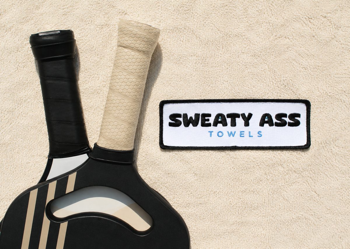 Sweaty Ass Towel in sand with crossed pickleball paddles